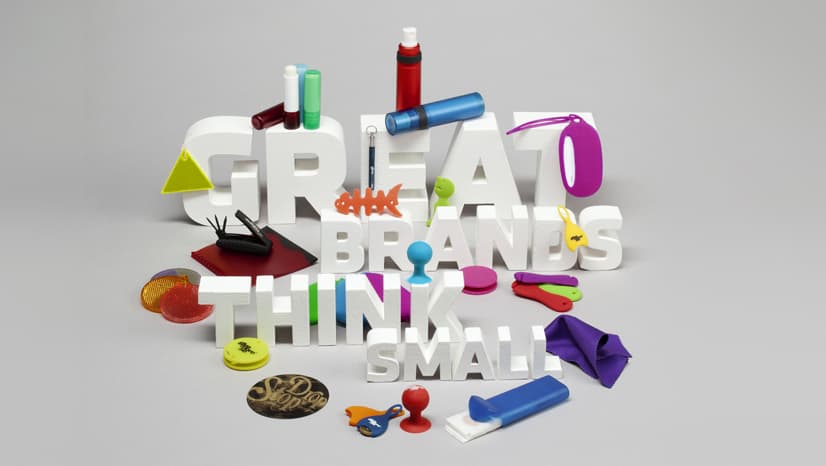Texten Great Brands Think Small i vita bokstäver omgiven av giveaways – reflexer, läppcerat, logoband och silikonprodukter